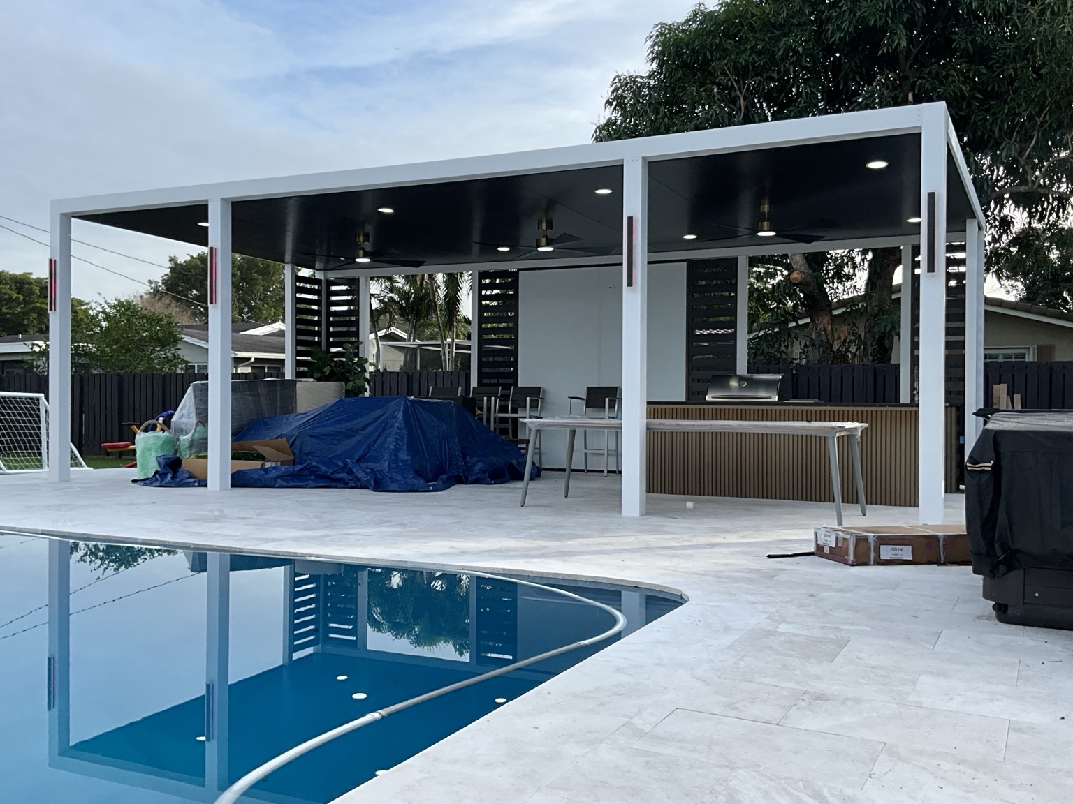 Completed white aluminum pergola with dark wood-look ceiling, fans, privacy panels, and outdoor kitchen beside a swimming pool
